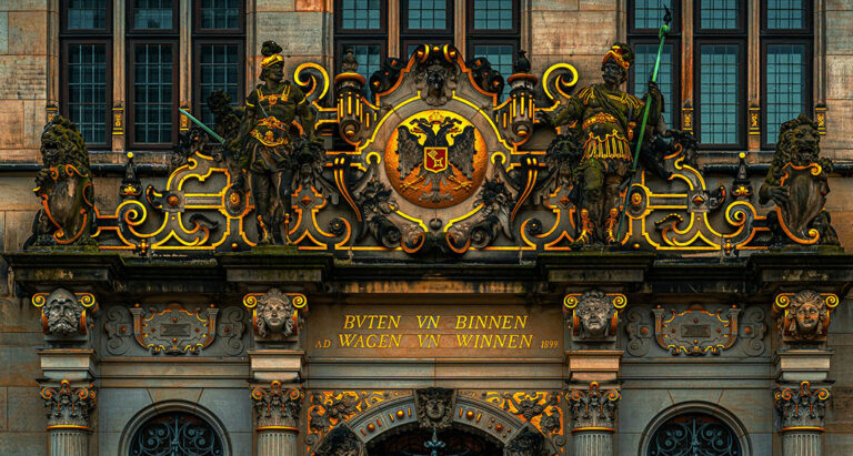 La photo montre le détail richement décoré d'un bâtiment historique, présentant des sculptures dorées, des armoiries, et des inscriptions, encadrées par des fenêtres et des colonnes classiques.