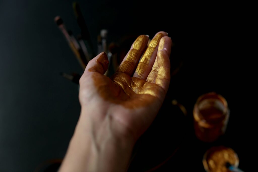 La photo montre une main ouverte avec la paume recouverte de peinture dorée, devant un fond sombre flou avec des pinceaux et des pots de peinture visibles en arrière-plan.
